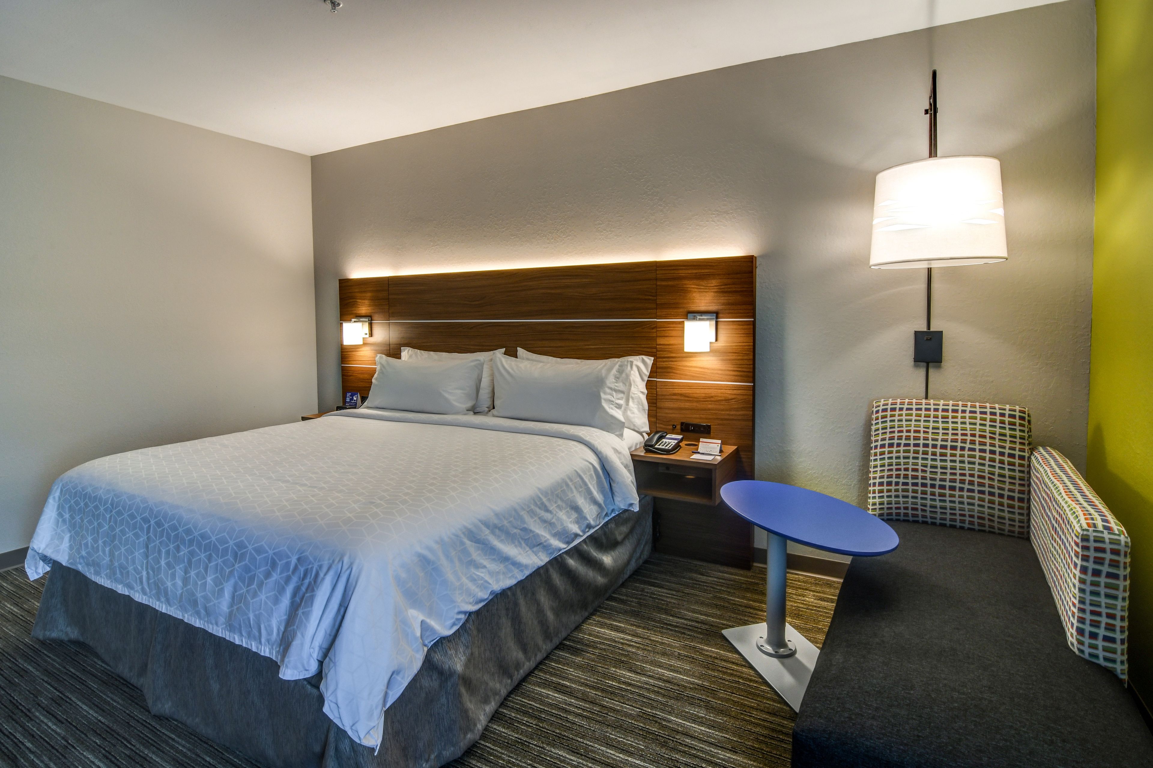 A hotel room with a large bed, a bedside table with a lamp, a small table, and a patterned bench.