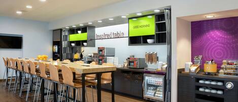 A modern dining area with a long table, chairs, a refrigerator, and a counter with food items.