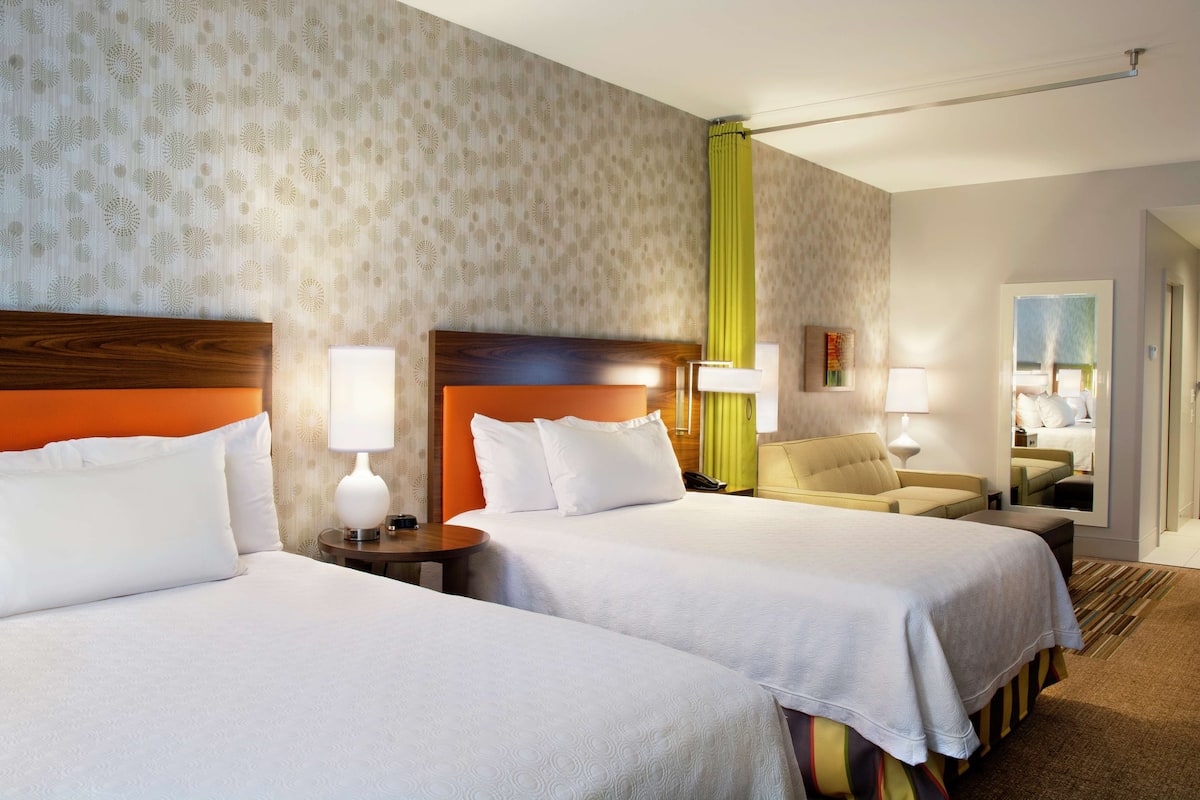 Une chambre d’hôtel avec deux lits, une tête de lit en bois, un rideau vert et un petit coin salon avec une chaise et une table.
