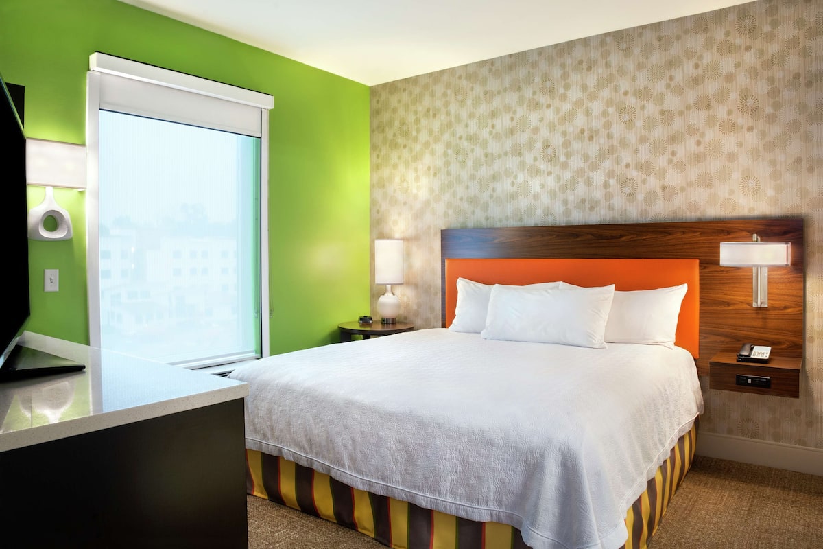 Une chambre d’hôtel avec un grand lit, un bureau avec un ordinateur portable, une fenêtre avec des stores et un papier peint à motifs sur le mur.
