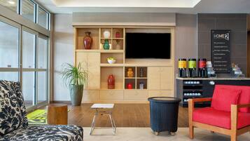 A modern hotel lobby with a large TV, a seating area with a patterned sofa, a small table, a potted plant, and a coffee station.