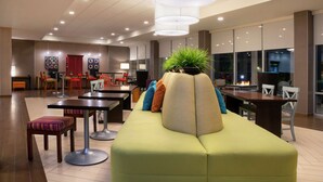 A modern lobby with a green sofa, dining tables, and chairs. There are potted plants and a fire feature in the background.