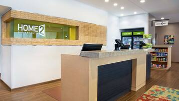 A hotel reception area with a reception desk, a sign reading "HOME 2 SUITES BY HILTON," and a refrigerator labeled "really cold" and "cold."