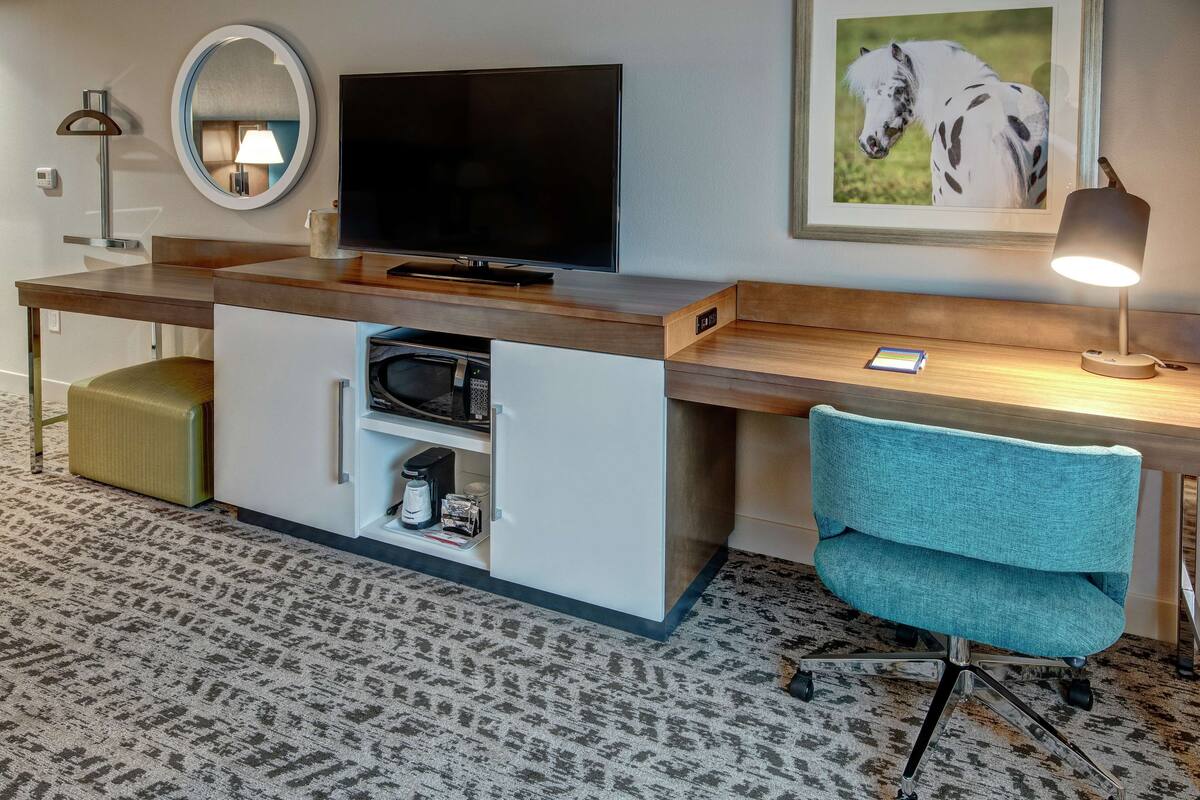 Hotelkamer met een houten bureau, een televisie, een magnetron, een koffiezetapparaat, een blauwgroene fauteuil, een lamp en een lijst met een afbeelding van een paard.