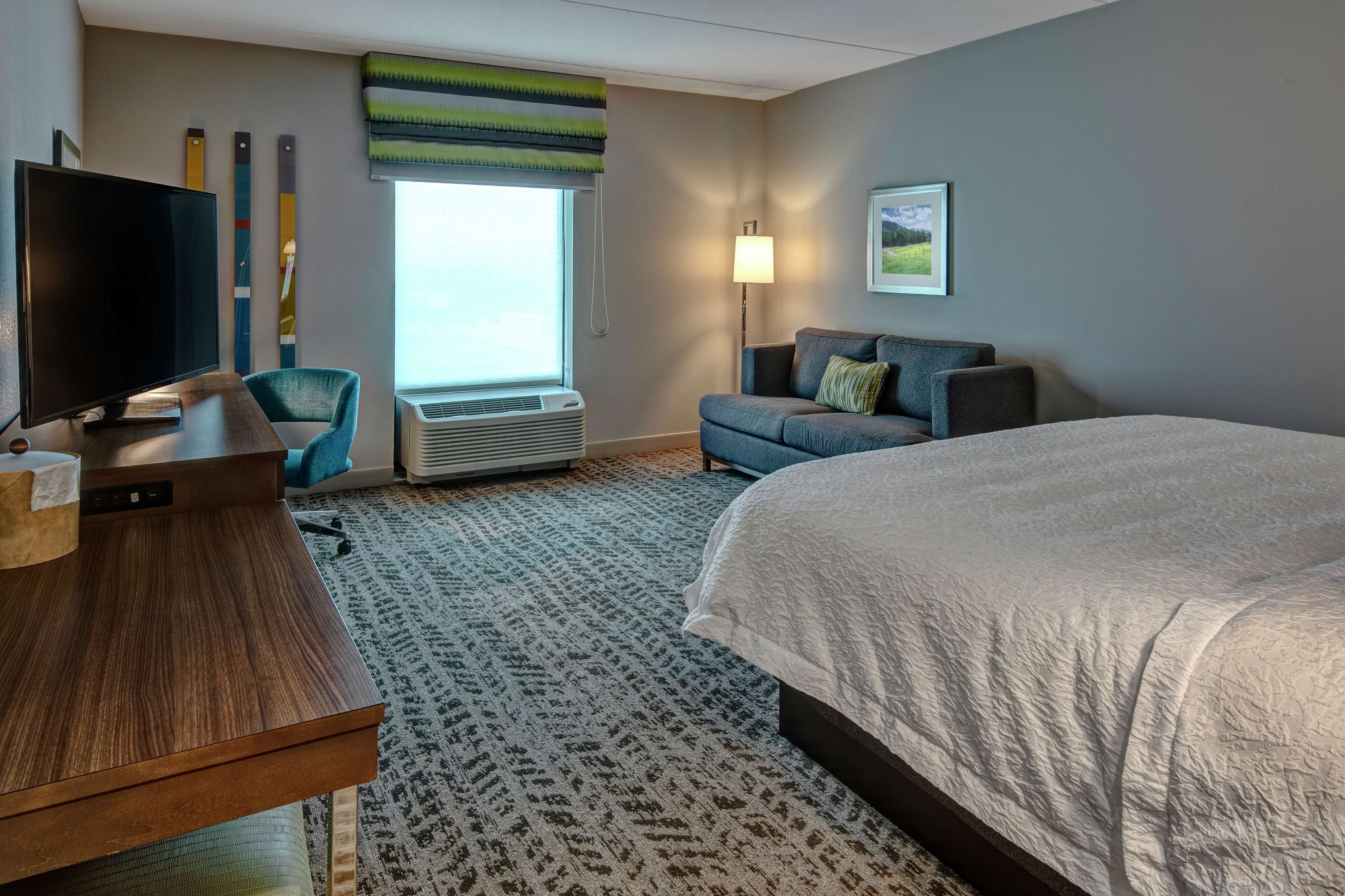 Ein Hotelzimmer mit einem großen Bett, einem Fernseher, einem Schreibtisch, einem Sessel, einer Couch, einer Lampe und einem Fenster mit Jalousien.