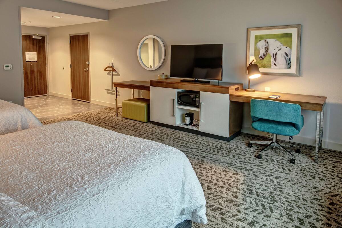 Een hotelkamer met een bed, een bureau met een computer, een televisie, een spiegel en een lijst met een afbeelding aan de muur.