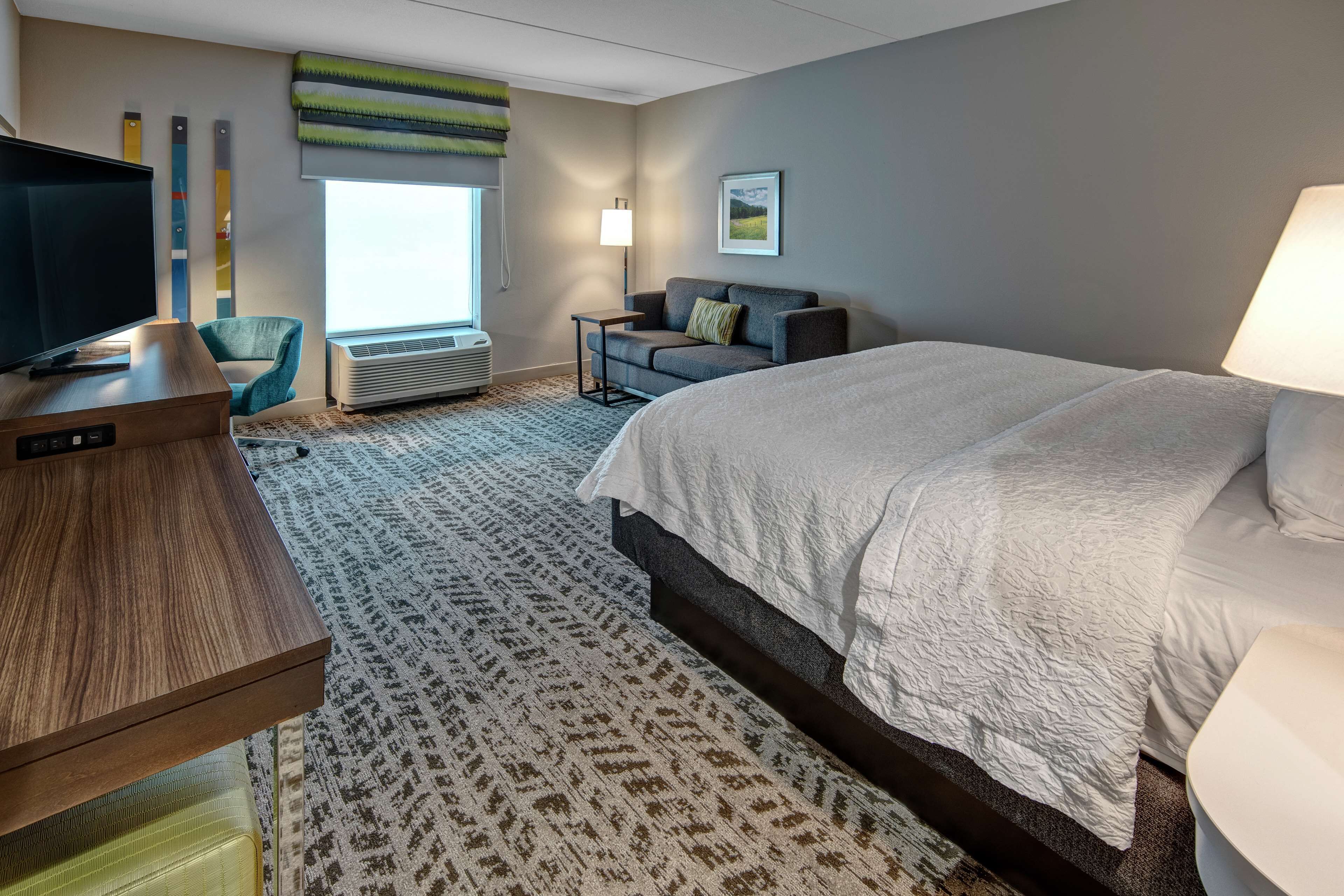 A hotel room with a bed, a TV, a desk, a chair, a lamp, and a window with blinds.