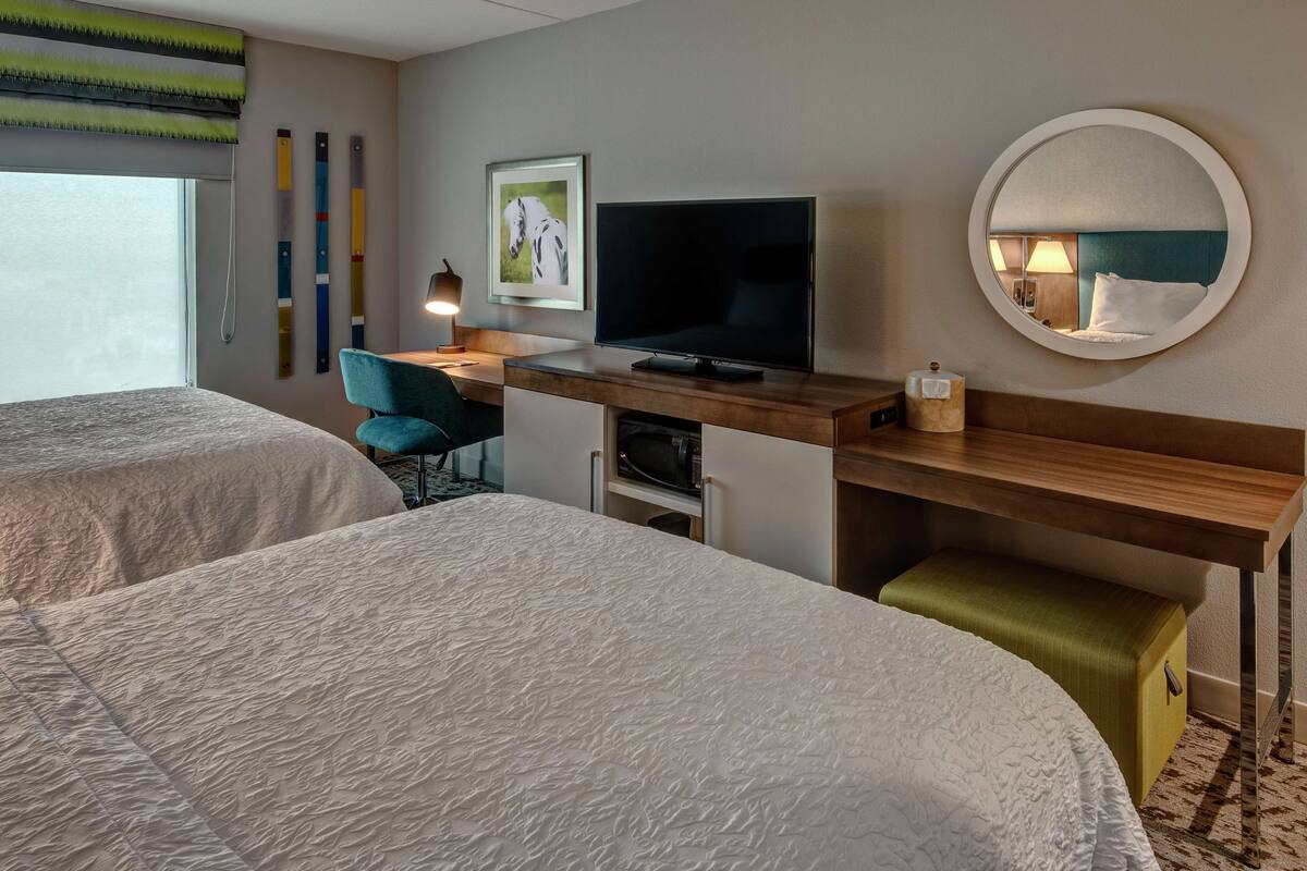 Een hotelkamer met twee bedden, een bureau met een televisie, een stoel, een spiegel en een raam met gordijn.