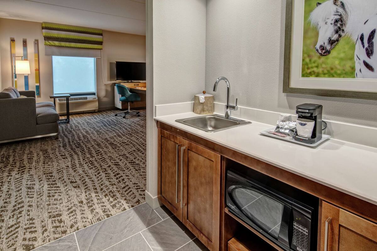 Een hotelkamer met een kitchenette, een bank, een bureau en een groot raam.