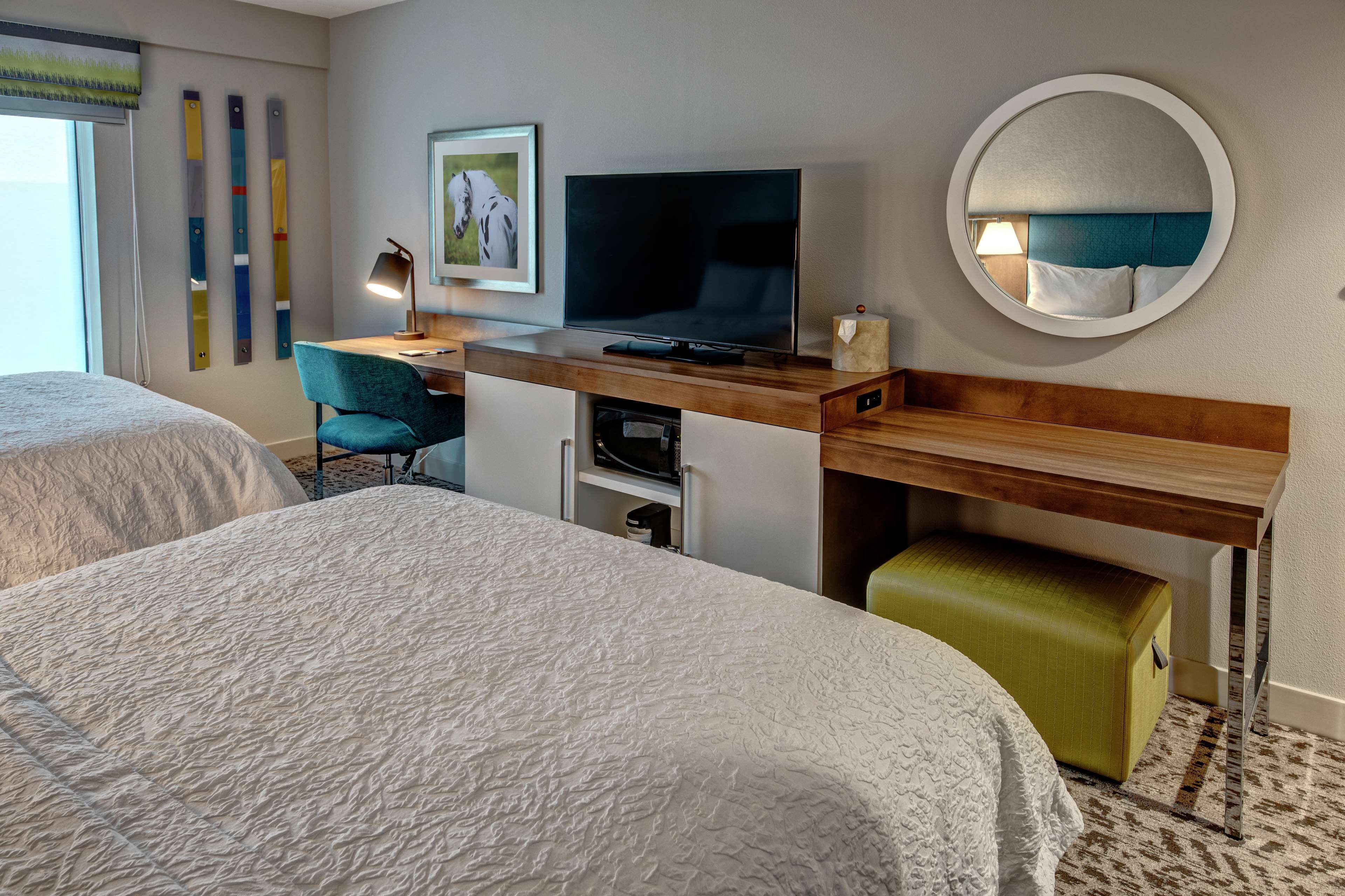 A hotel room with two beds, a desk with a television, a mirror, and a green ottoman.