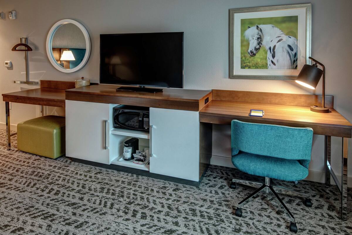 Een hotelkamer met een houten bureau, een televisie, een lijst met een afbeelding van een paard, een blauwgroene stoel en een magnetron.