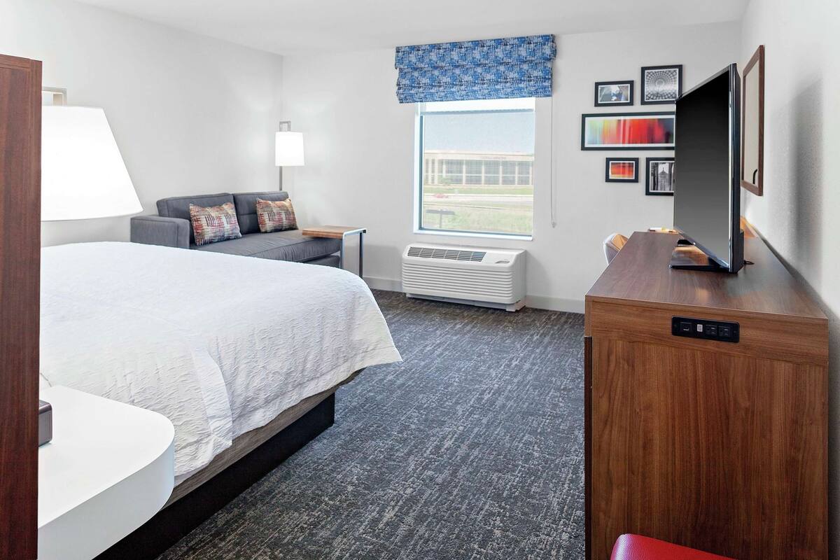 A hotel room with a bed, a desk, a TV, a window with a view, and a wall with framed pictures.