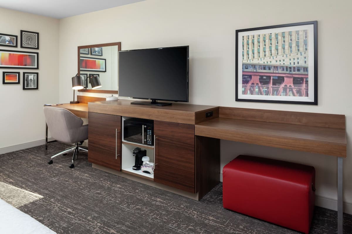A modern hotel room with a wooden desk, a flat-screen TV, a red ottoman, and a framed cityscape picture on the wall.