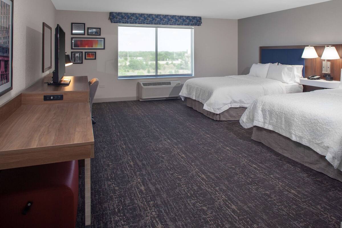 A hotel room with two beds, a desk, a TV, and a large window.