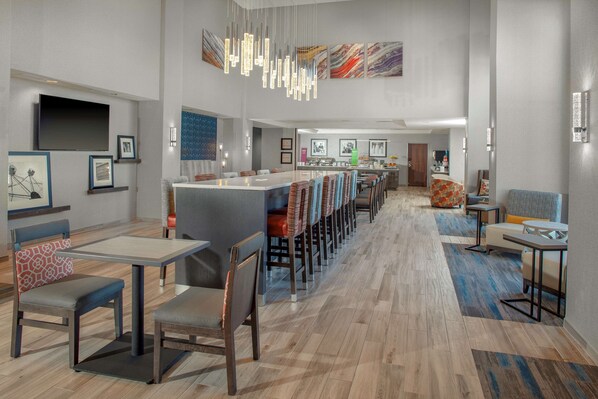 A modern hotel lobby with a long dining area, multiple seating options, and a large TV screen.