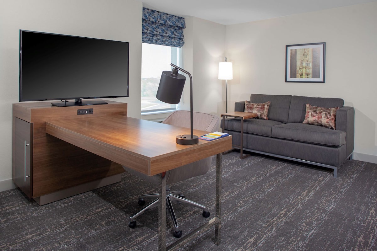A modern hotel room with a wooden desk, a flat-screen TV, a grey sofa, and a framed picture on the wall.