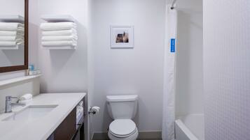 Un baño moderno con un lavamanos, espejo y retrete blancos. Hay toallas apiladas en un estante y una fotografía enmarcada en la pared.
