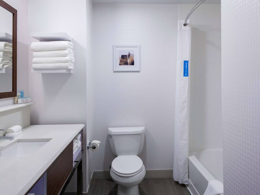 Une salle de bain moderne avec un lavabo blanc, un miroir et des toilettes. Des serviettes sont pliées sur une étagère, et une photo encadrée est accrochée au mur.