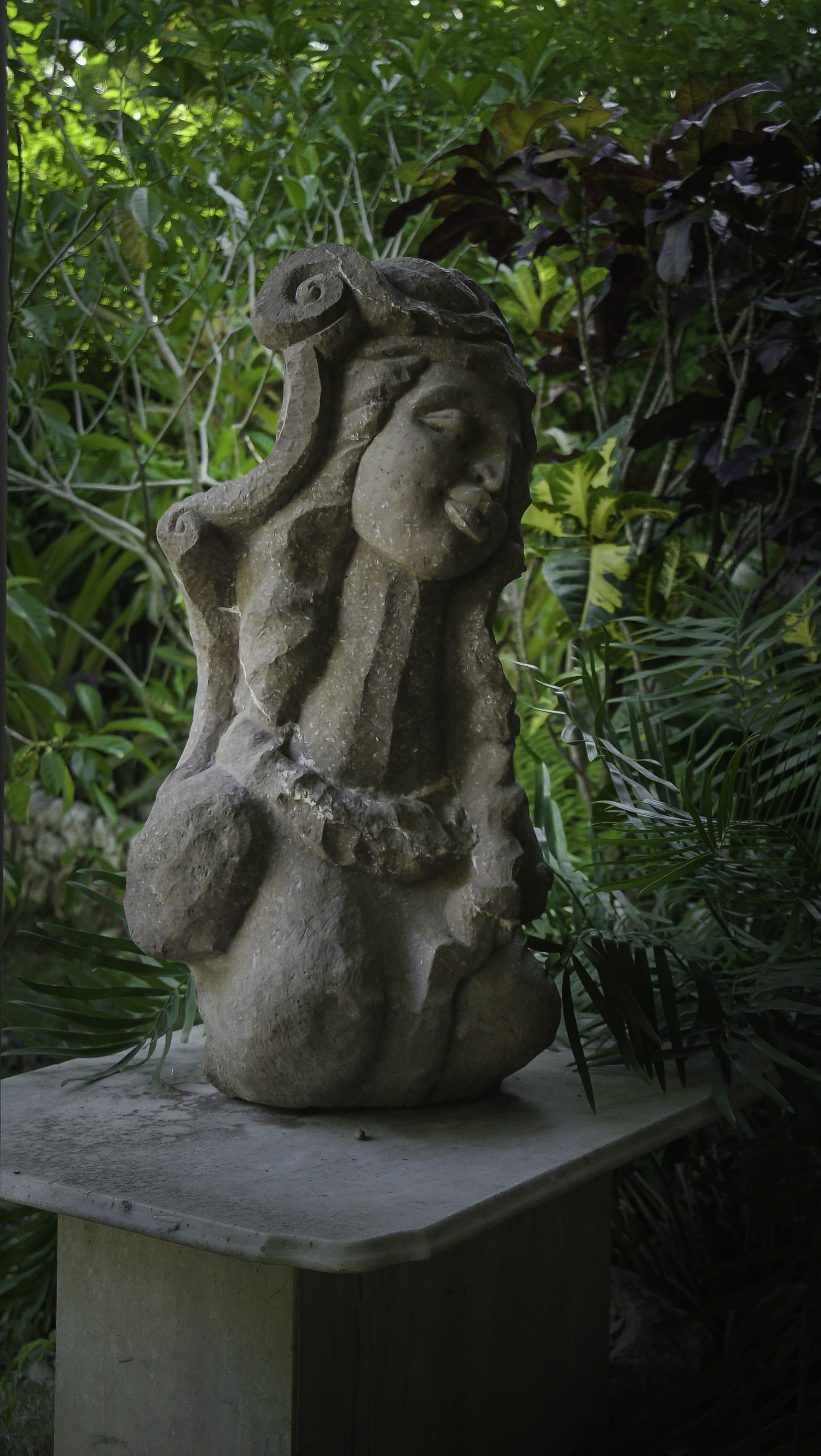 Una statua in pietra raffigurante una donna con un’aureola, posata su un piedistallo in cemento in un giardino con fitta vegetazione sullo sfondo.