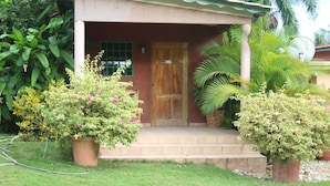 A small house with a wooden door and a porch. There are potted plants and a garden in front. - Le Jardin Hotel Restaurant (Jacmel)
