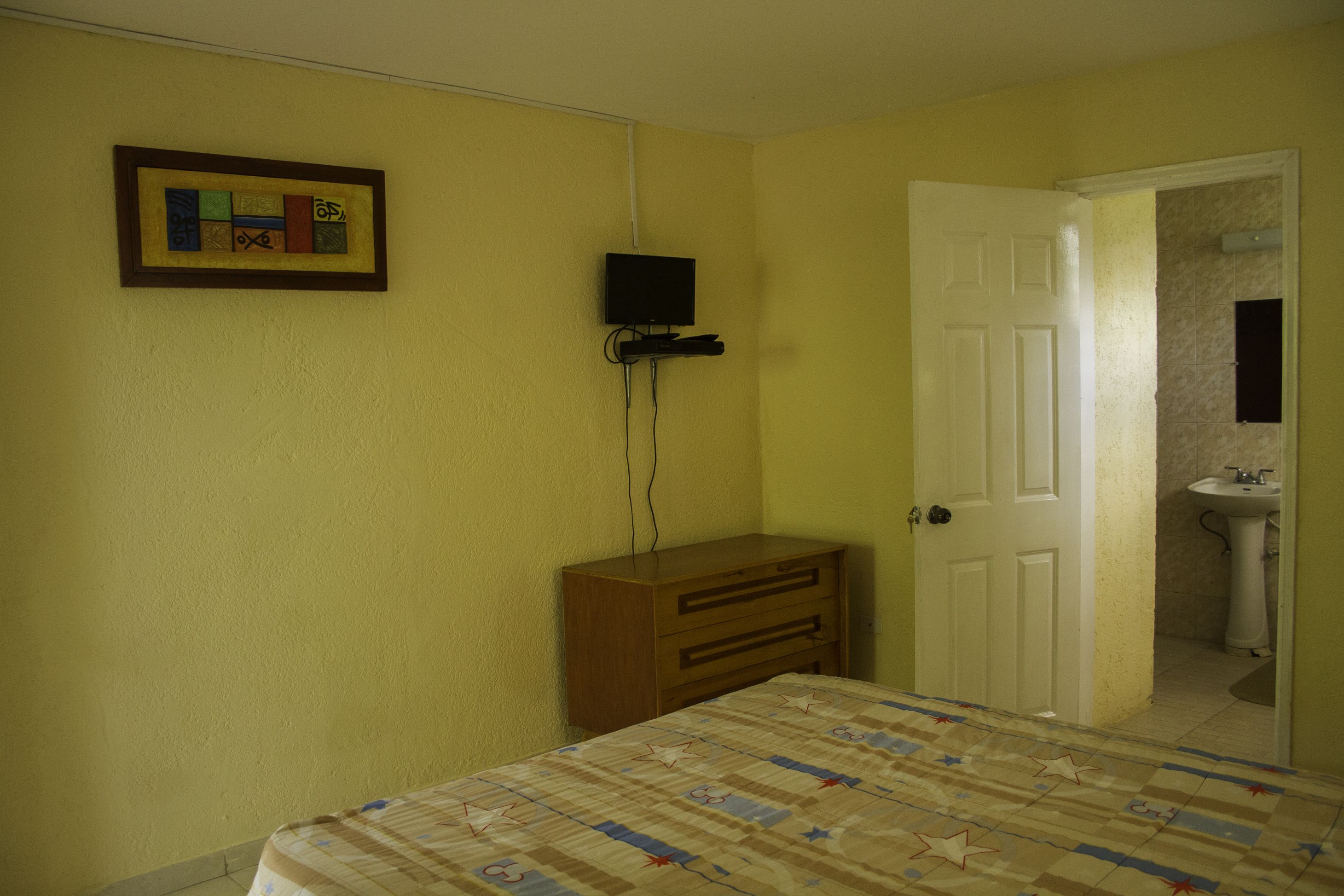 A bedroom with a bed, a dresser, a television mounted on the wall, and a door leading to a bathroom.