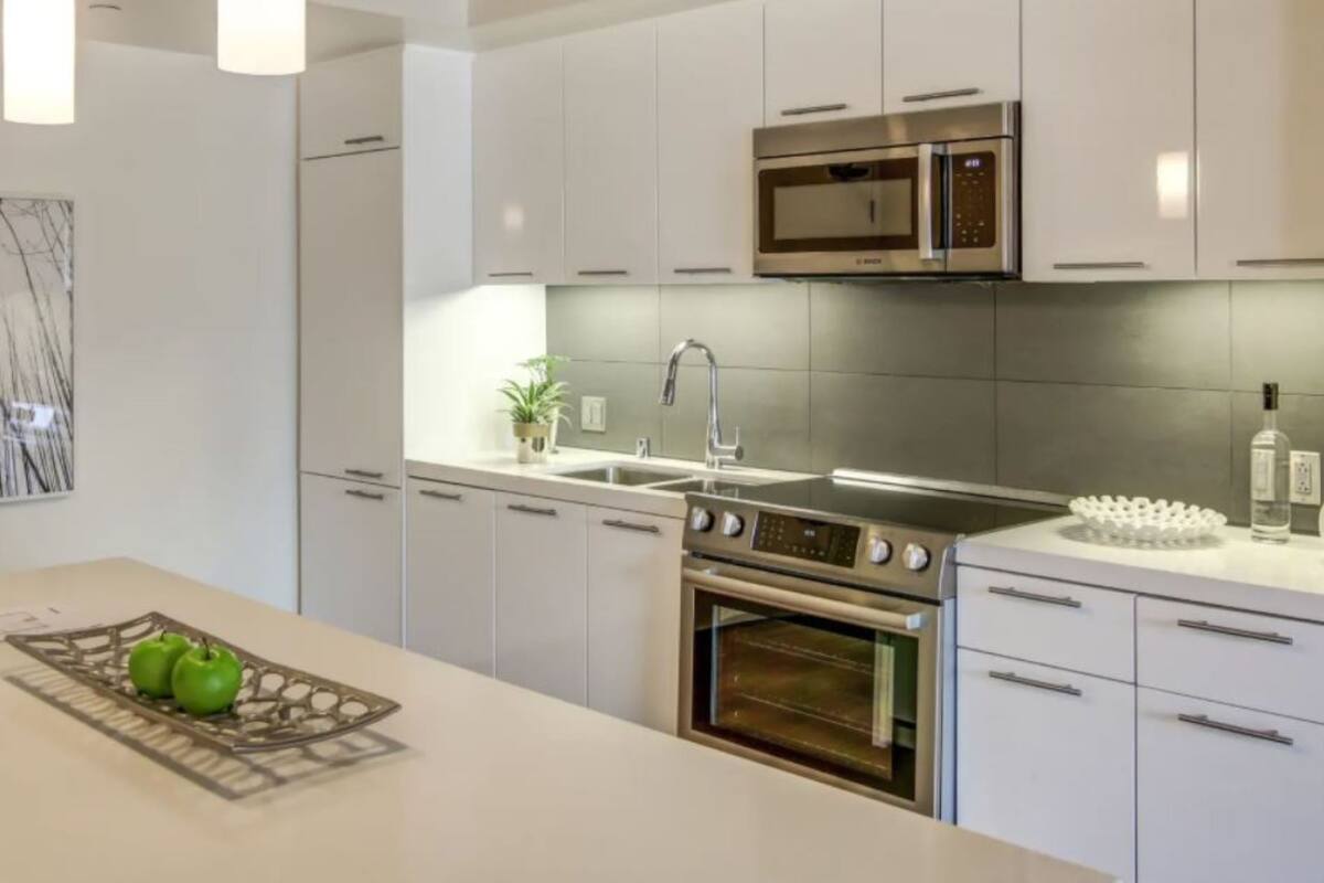 A modern kitchen with white cabinetry, a stainless steel oven, a microwave, and a large island with a tray of green apples.