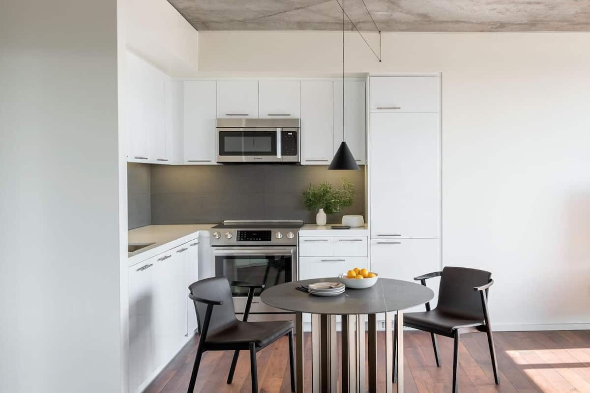 A modern kitchen with white cabinetry, a built-in microwave, and a dining area with a round table and two chairs.