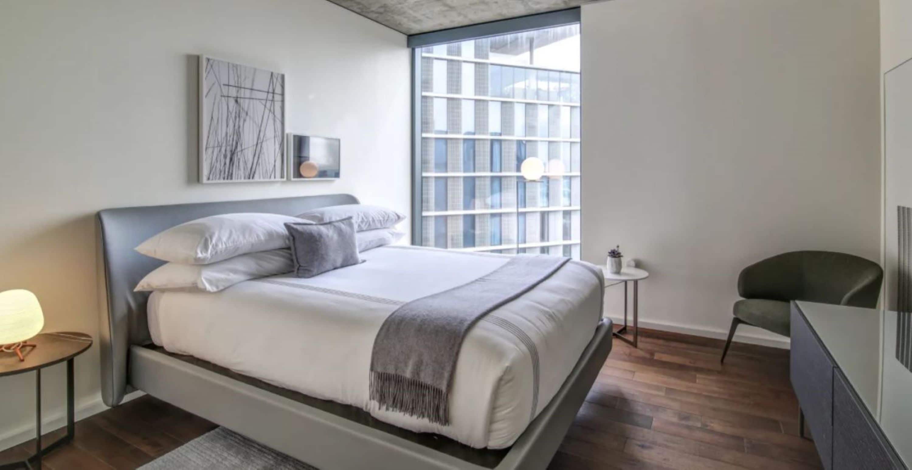 Camera moderna con un letto grande, un comodino, una sedia e una finestra con vista sulla città.