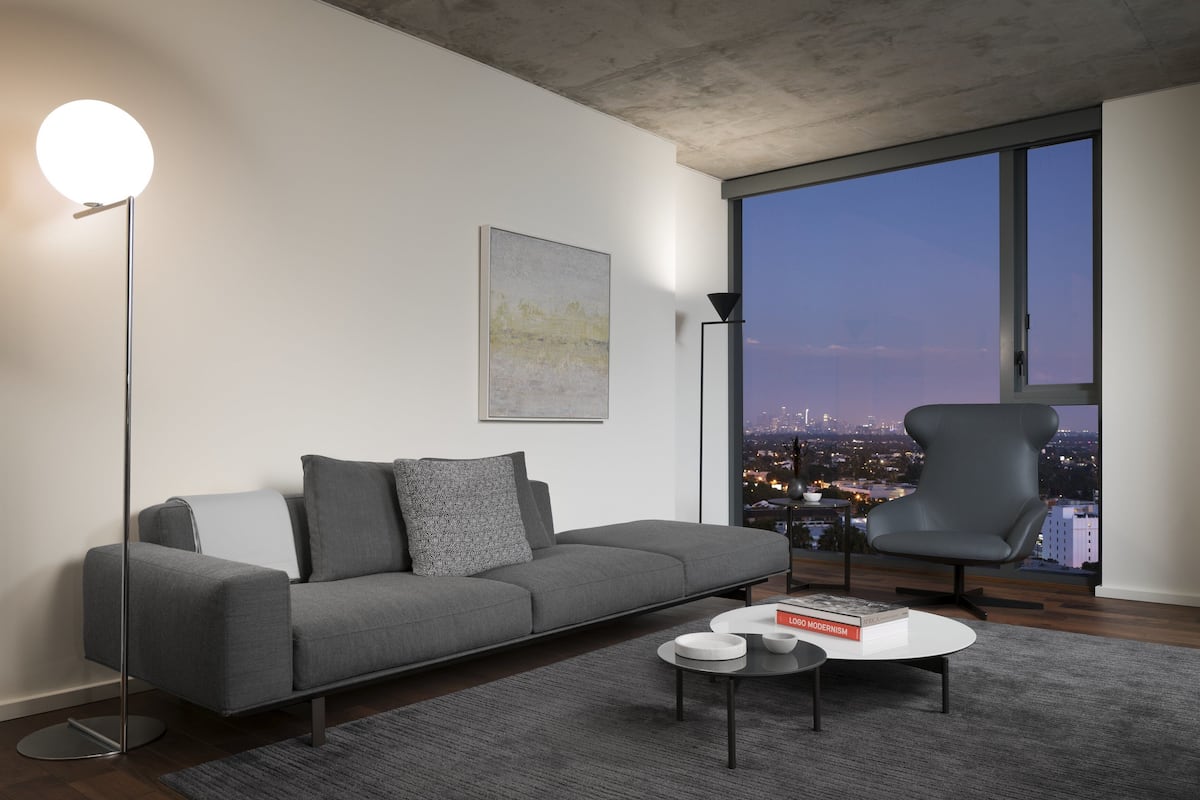 A modern living room with a grey sofa, a round coffee table, and a floor lamp. A large window offers a view of a cityscape at dusk.