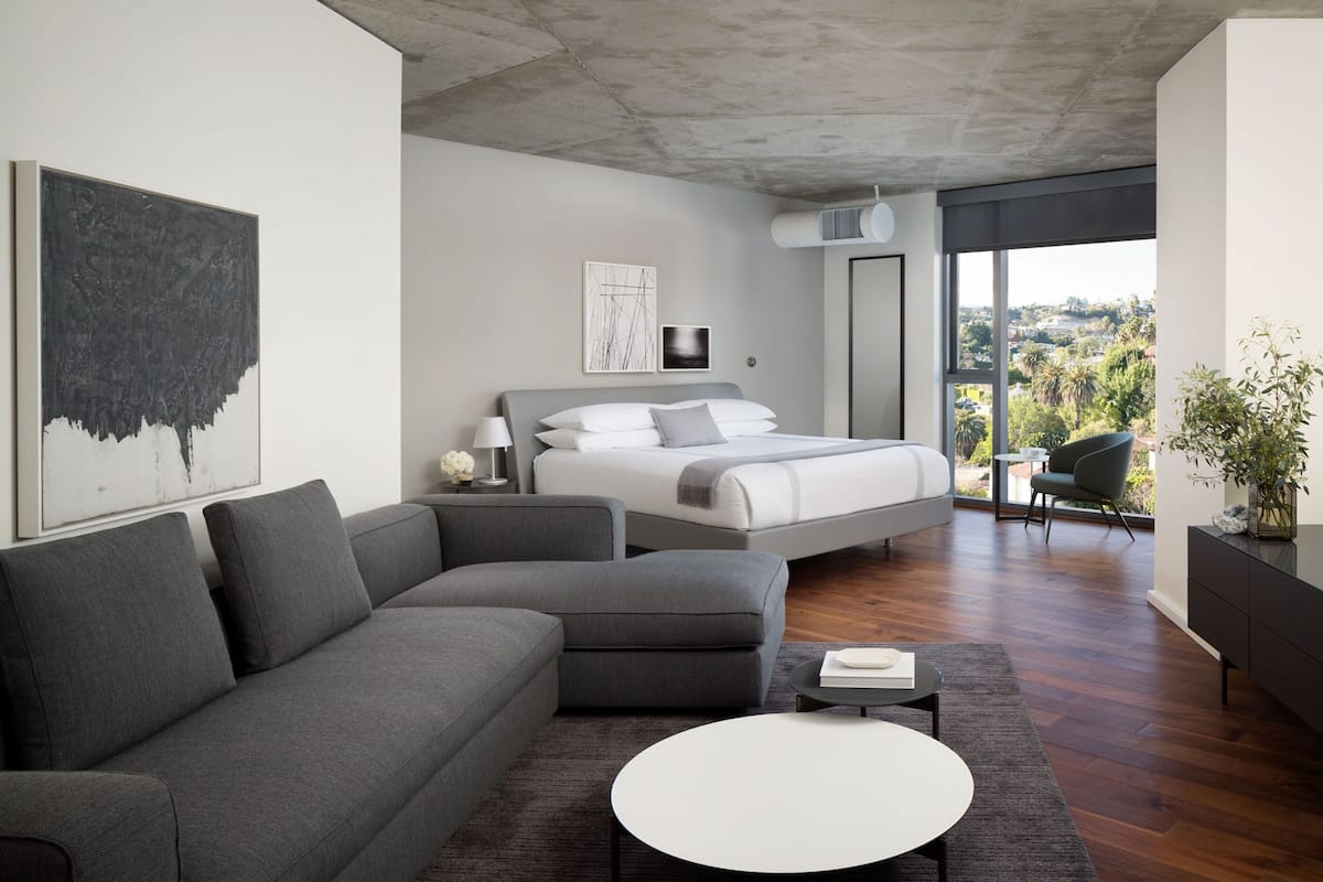 A modern bedroom with a large bed, a gray sofa, a round coffee table, and a view of the city.