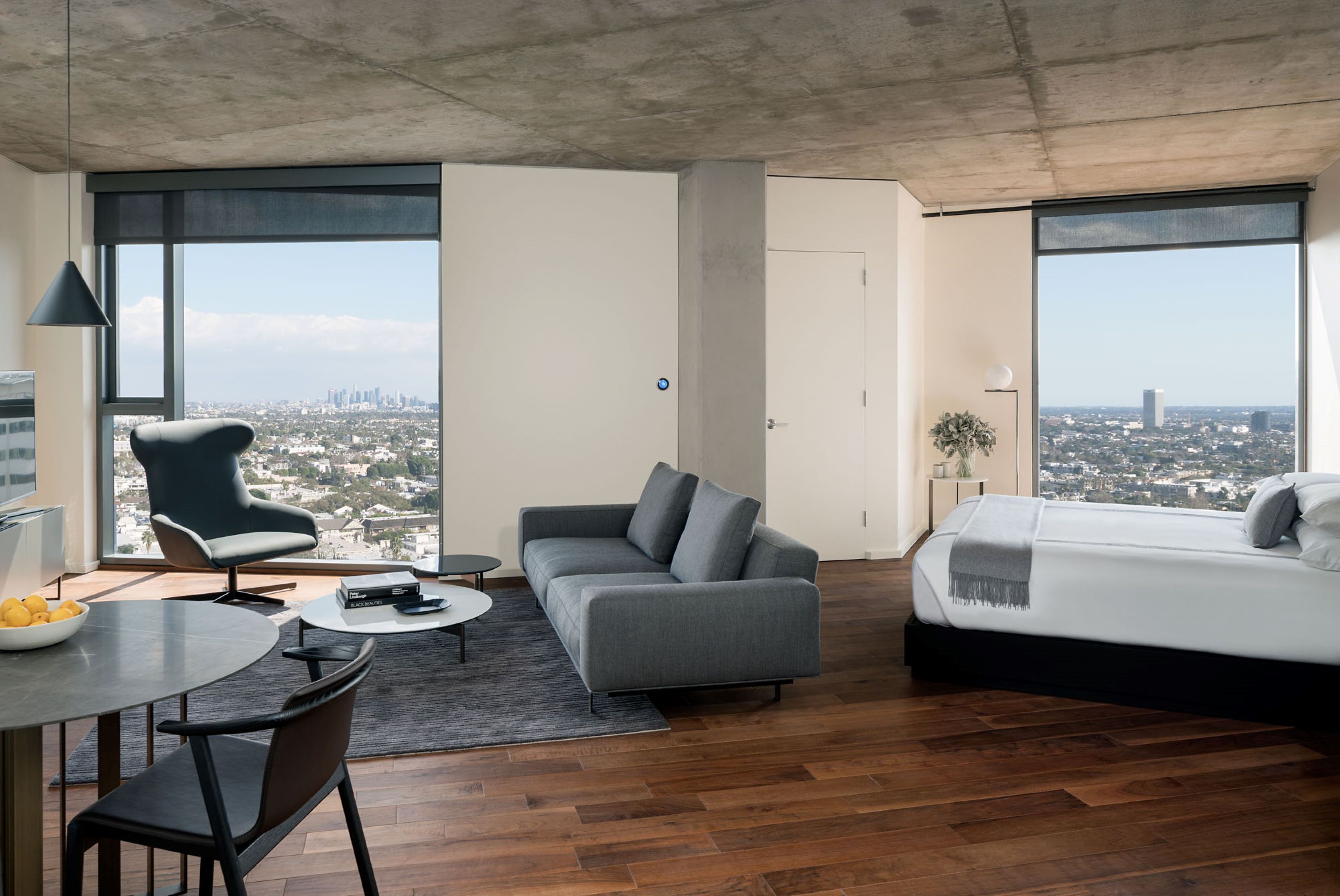 A modern hotel room with a large bed, a sofa, a dining area with a table and chairs, and large windows offering city views.