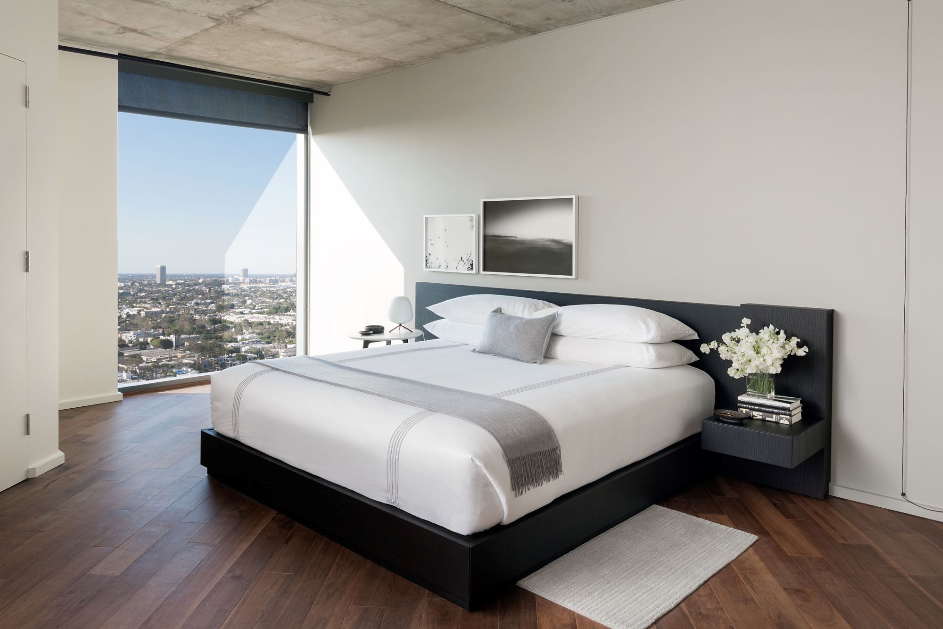 A modern bedroom with a large bed, a nightstand, and a view of a cityscape through a large window.