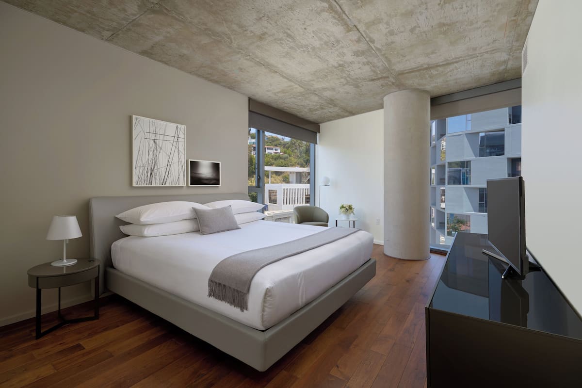 A modern bedroom with a large bed, a bedside table with a lamp, a TV, and a view of buildings through large windows.