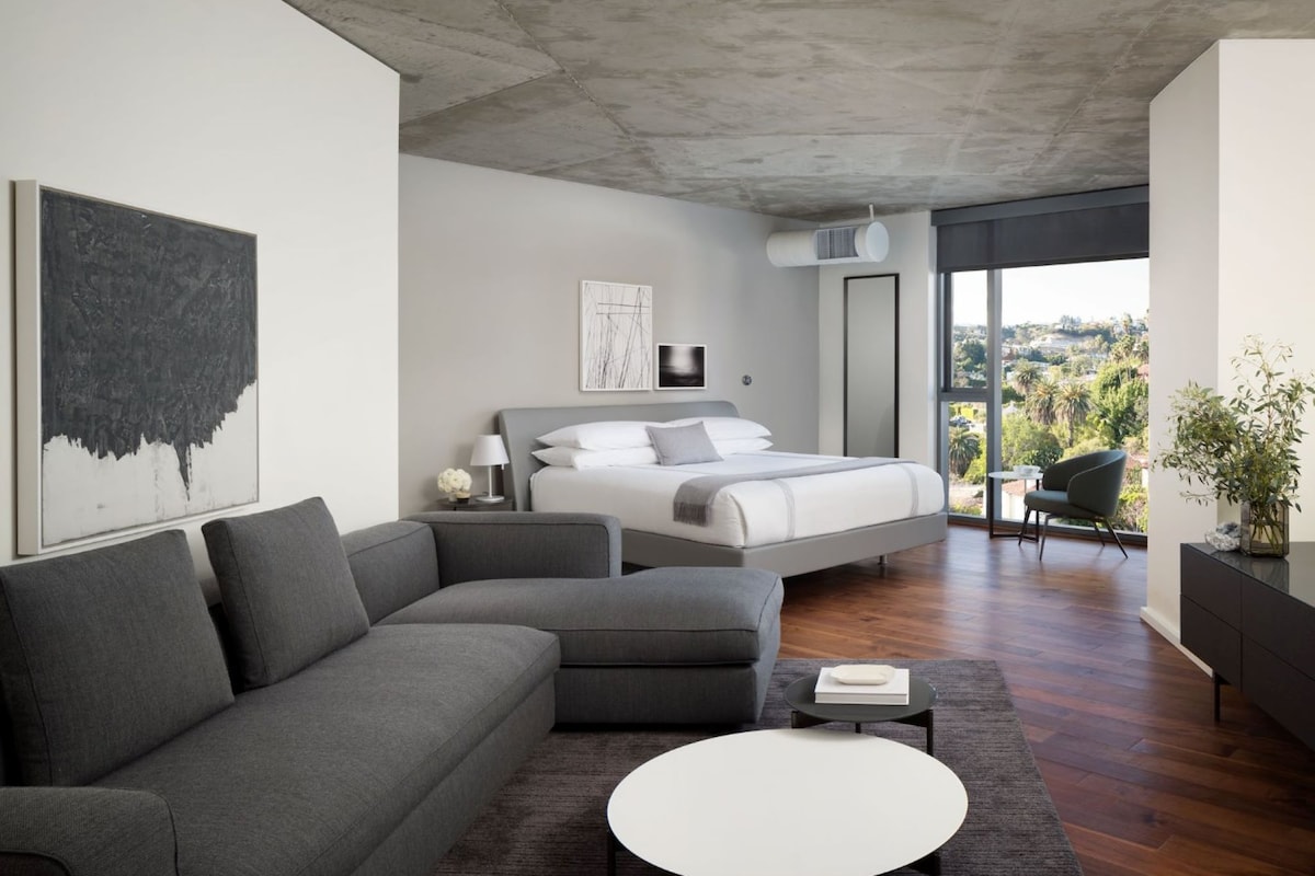 Un dormitorio moderno con una cama grande, un sofá gris, una mesita redonda y vistas al exterior.
