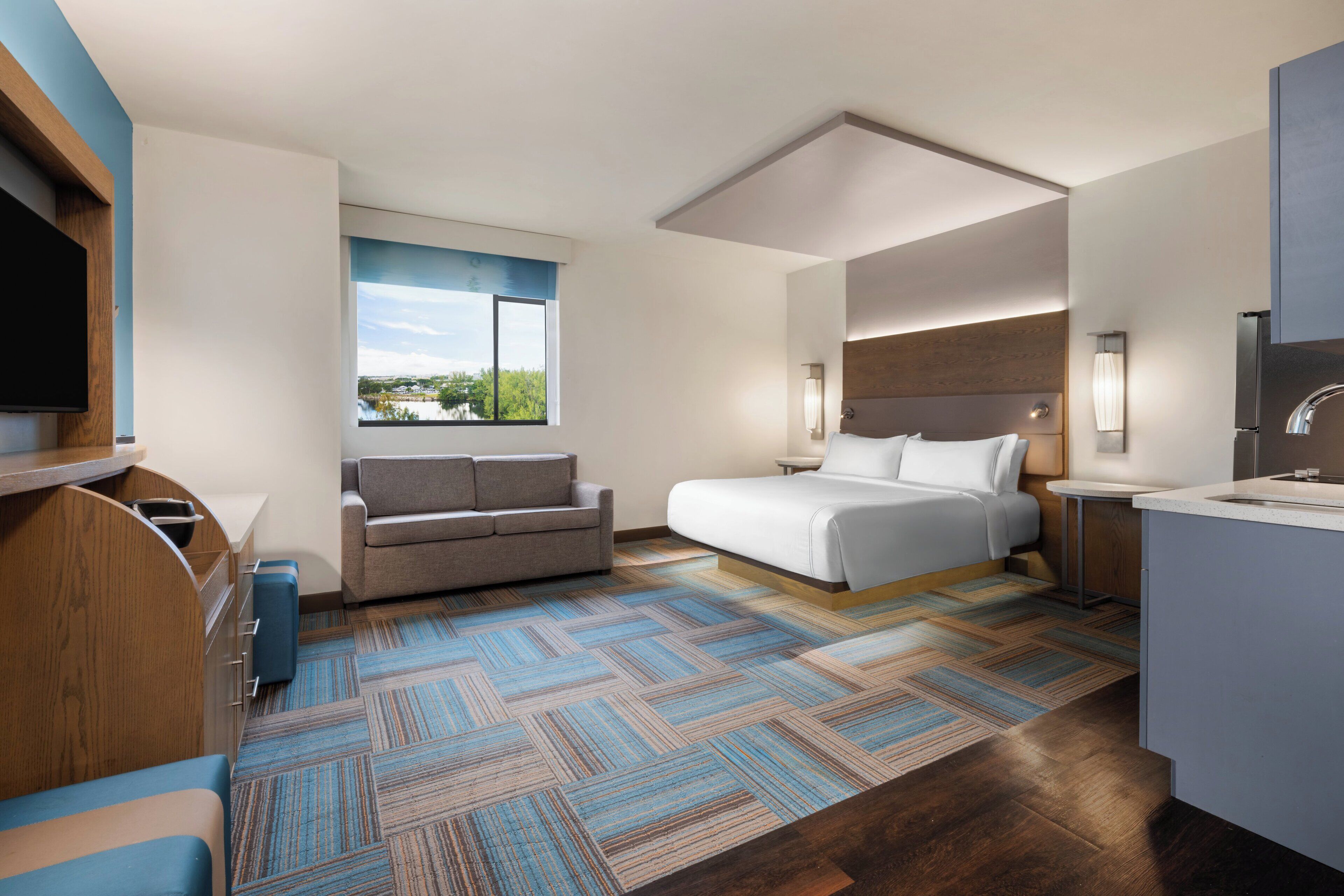 A modern hotel room with a bed, a sofa, a desk, a TV, and a view of the outdoors.