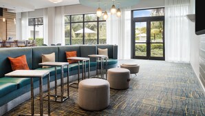 A modern lounge area with a teal sofa, round ottomans, and metal tables. Large windows offer a view of palm trees and a white umbrella. - EVEN Hotel Miami - Airport by IHG (Miami)