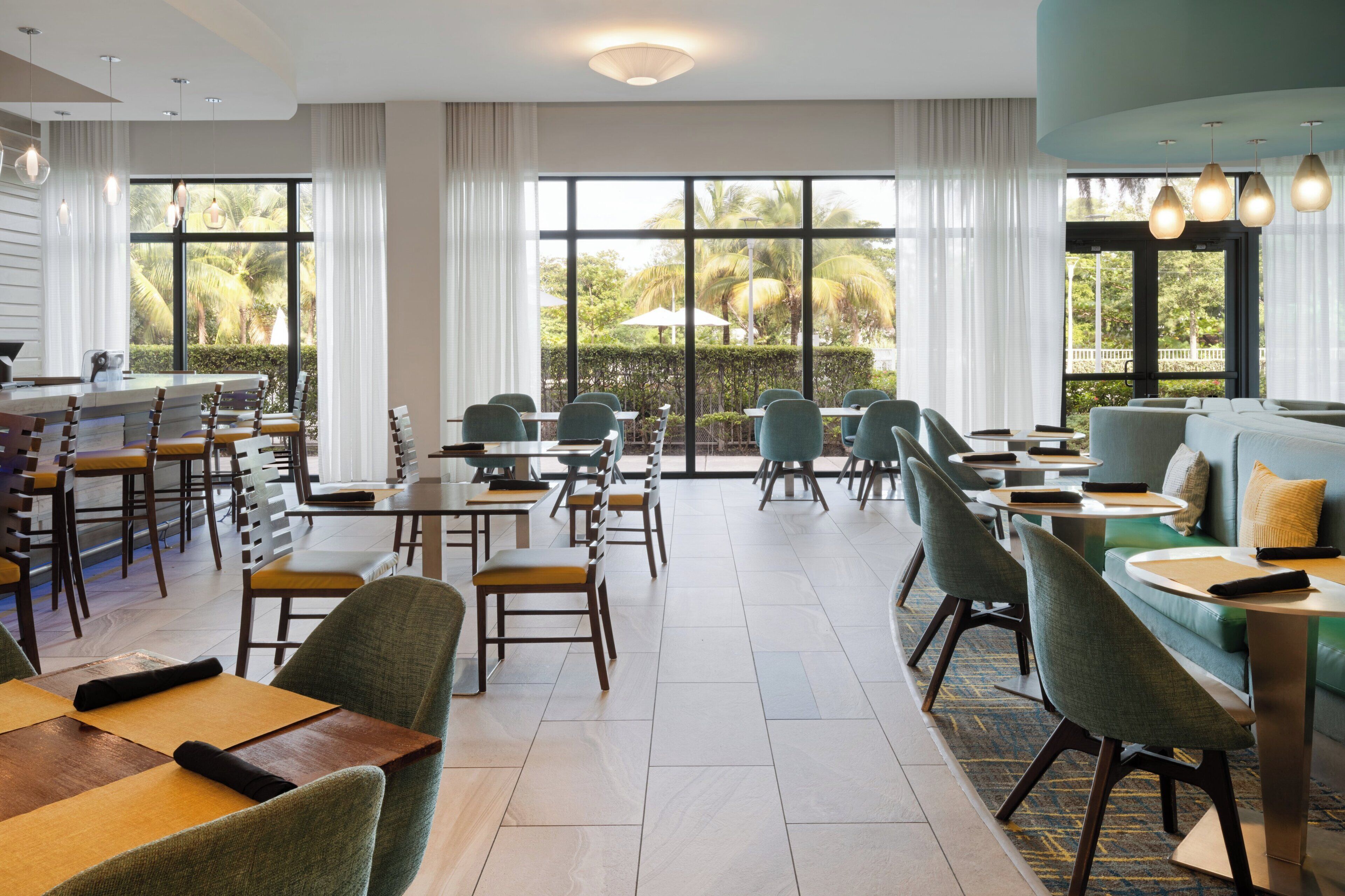Un comedor de hotel con varias mesas y sillas, ventanas amplias con cortinas y vistas a zonas verdes.