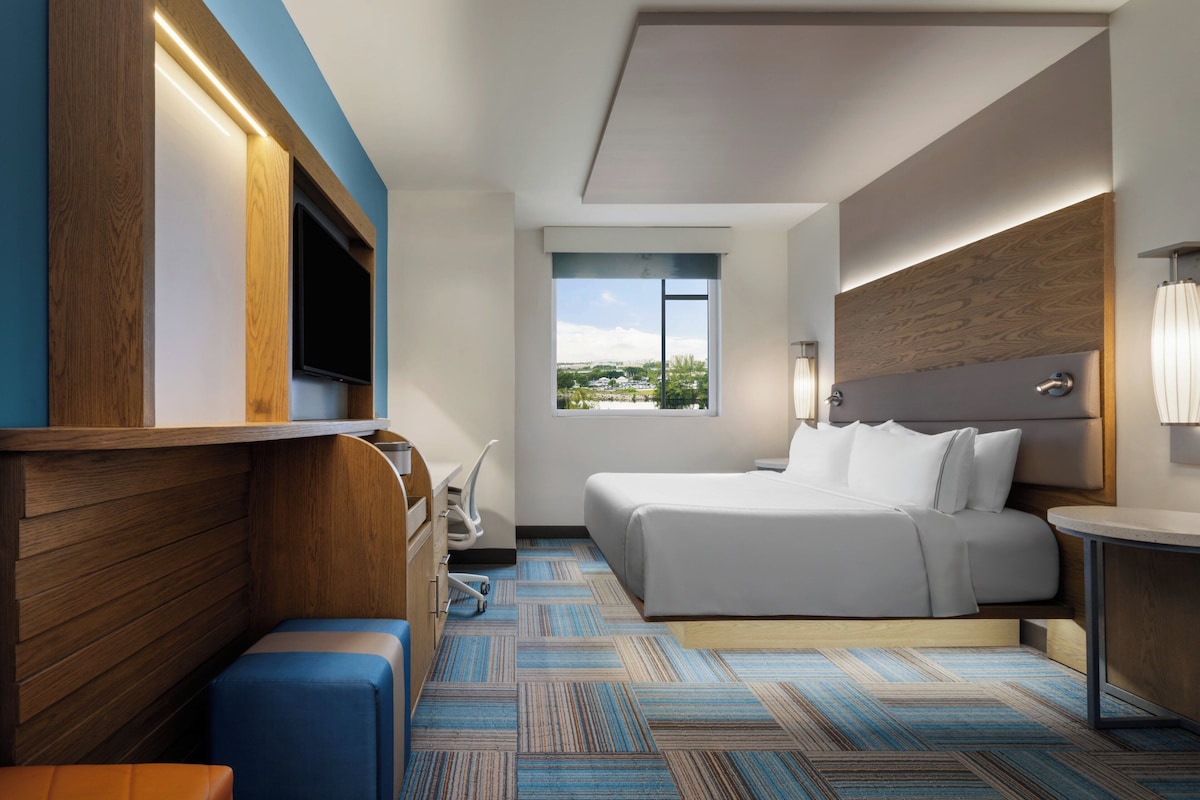 Una habitación de hotel moderna con una cama grande, un televisor montado en la pared, un ventanal con vista a la ciudad y un cabecero de madera.