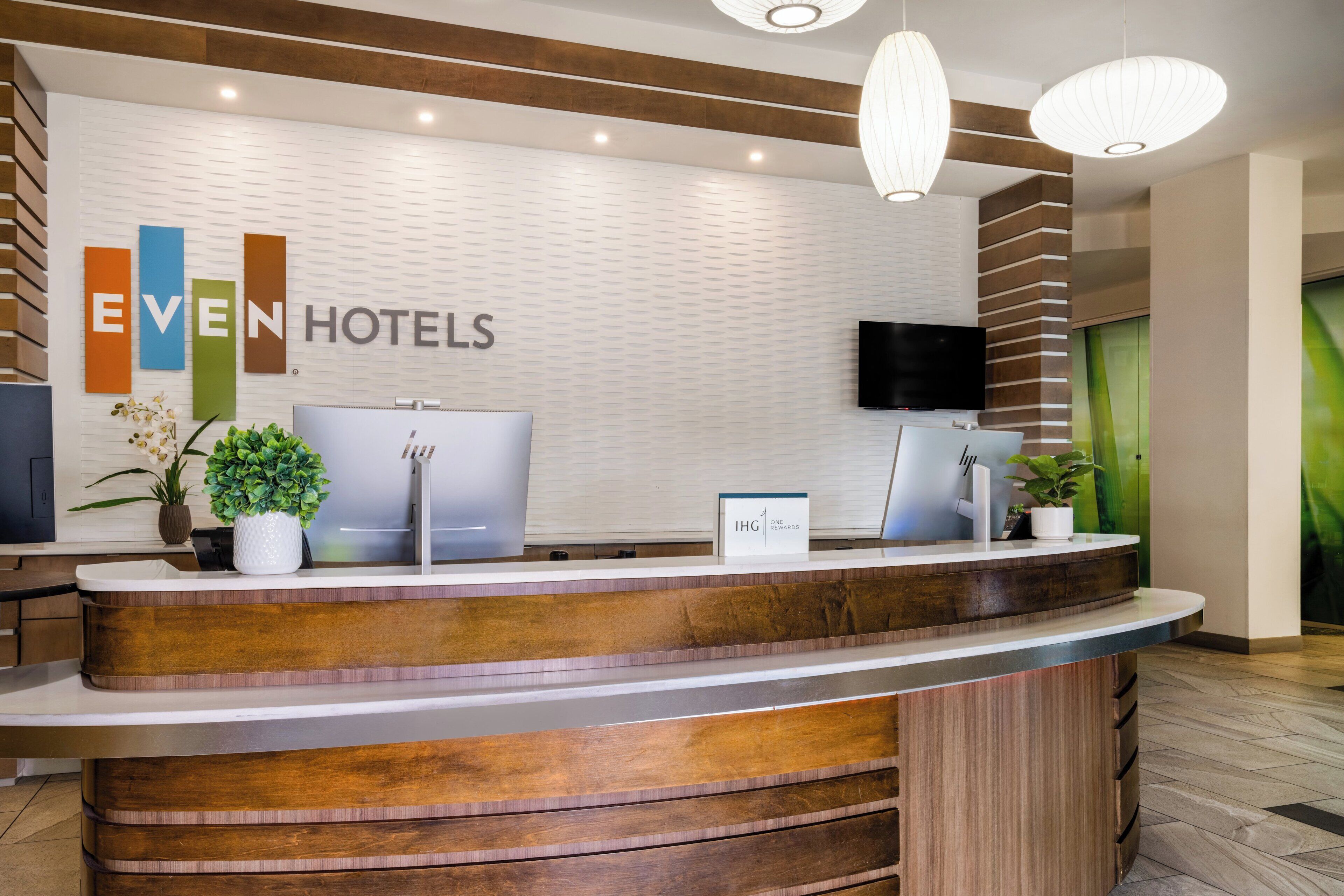 Un mostrador de recepción de diseño moderno, un letrero de Even Hotels, un televisor de pantalla plana y una planta en maceta.