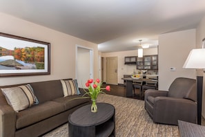 A living room with a sofa, a coffee table with flowers, a dining area with a table and chairs, and a kitchen in the background. - Candlewood Suites Bensalem - Philadelphia Area by IHG (Feasterville-Trevose)