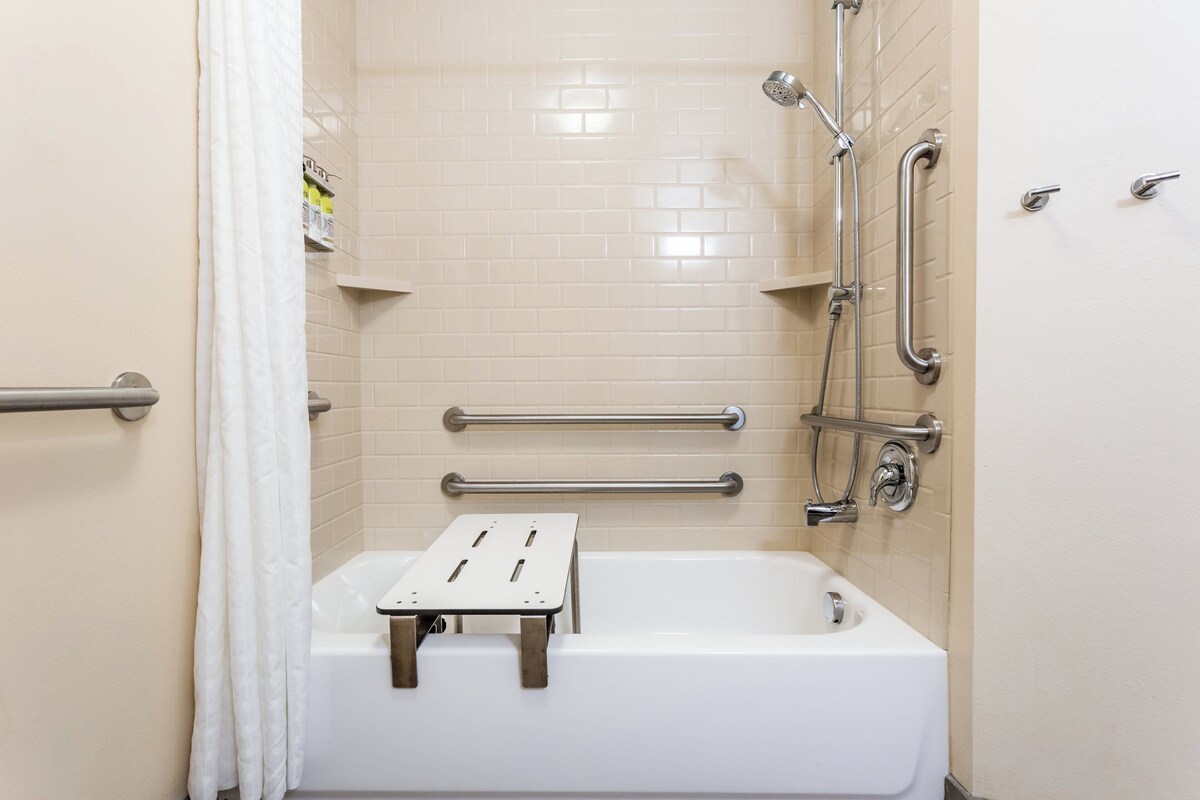 A bathroom with a white bathtub, grab bars, a shower head, and a toilet.