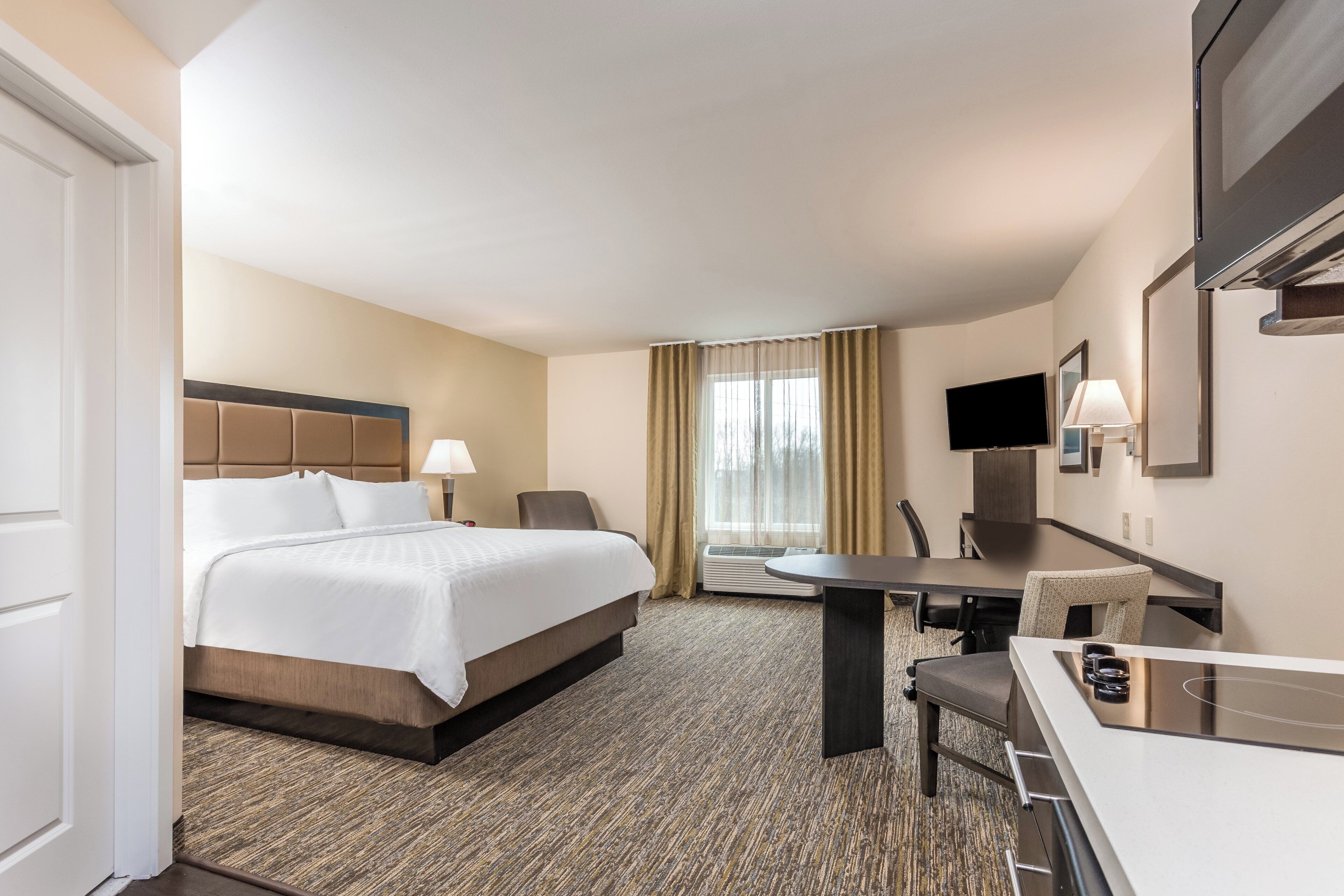 Quarto de hotel com uma cama, escrivaninha, cadeira, televisão e janela com cortinas.