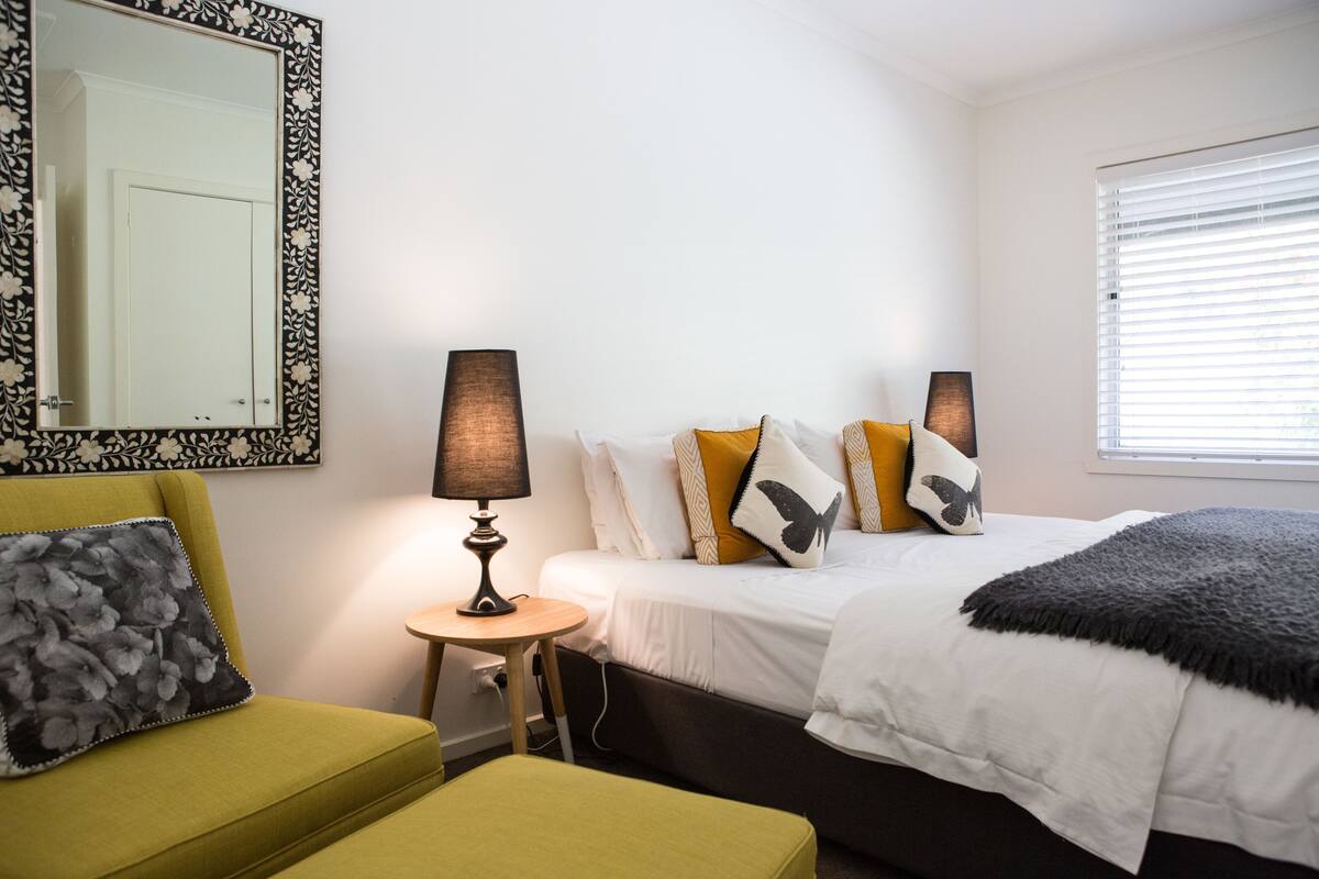 A hotel room with a bed, a small table with a lamp, a chair, and a large mirror on the wall.