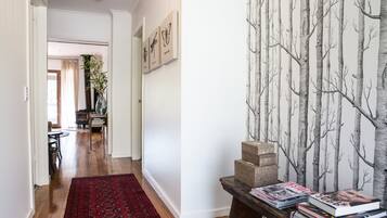 Un pasillo con piso de madera, un tapete rojo estampado, una mesa con libros y una revista, y una pared con papel tapiz de ramas de árbol.
