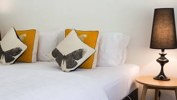 Una cama bien hecha con ropa de cama blanca y almohadillas decorativas, una mesita de madera redonda con una lámpara, y una pared lisa al fondo.