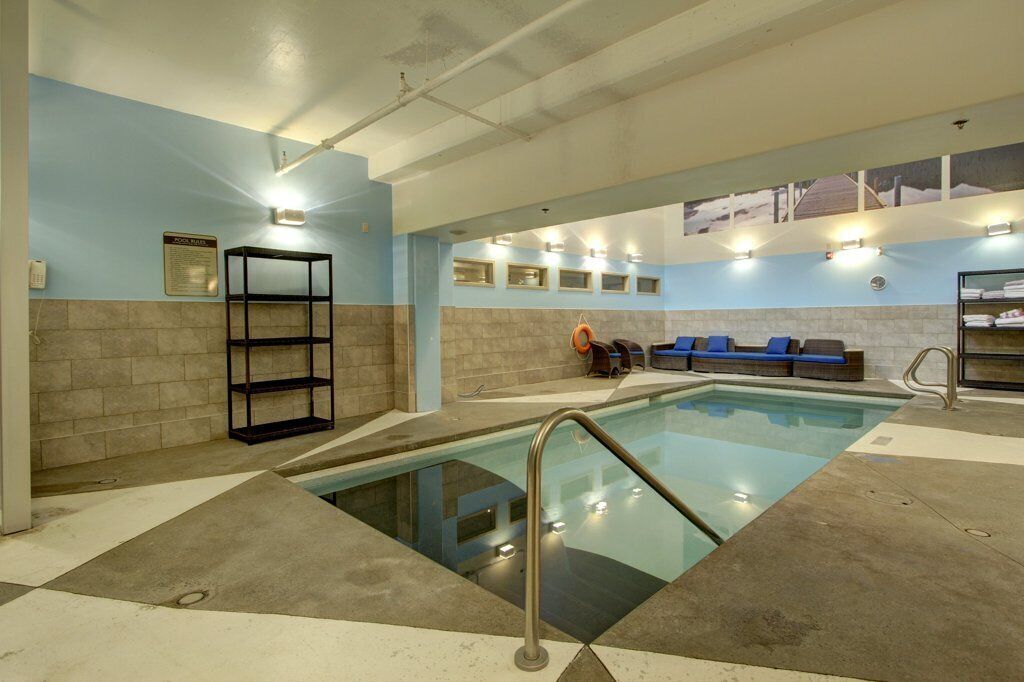 Une piscine intérieure avec un sol carrelé, un escalier métallique et des sièges rembourrés bleus le long du mur.