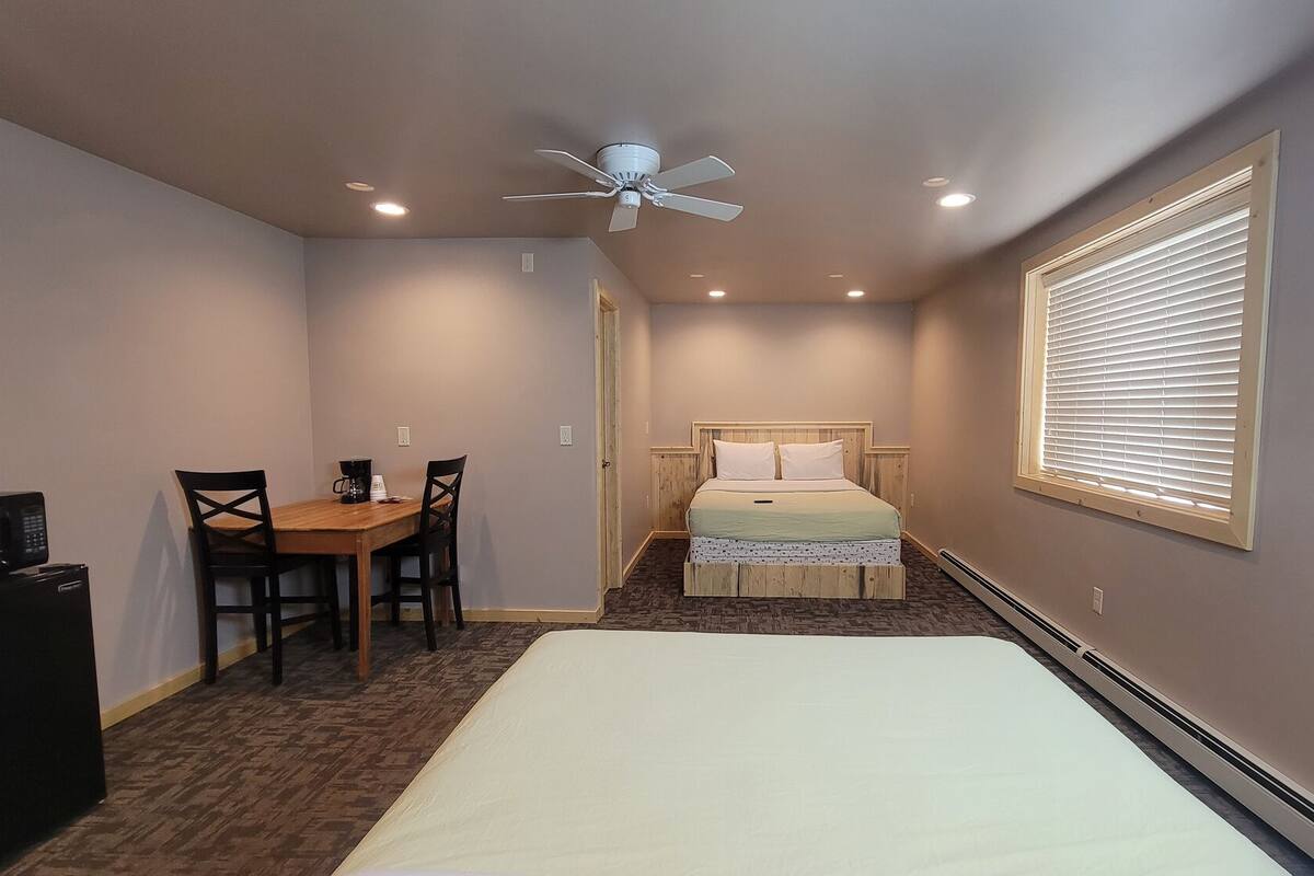 A compact bedroom with a bed, a small table with chairs, a ceiling fan, and a window with blinds.