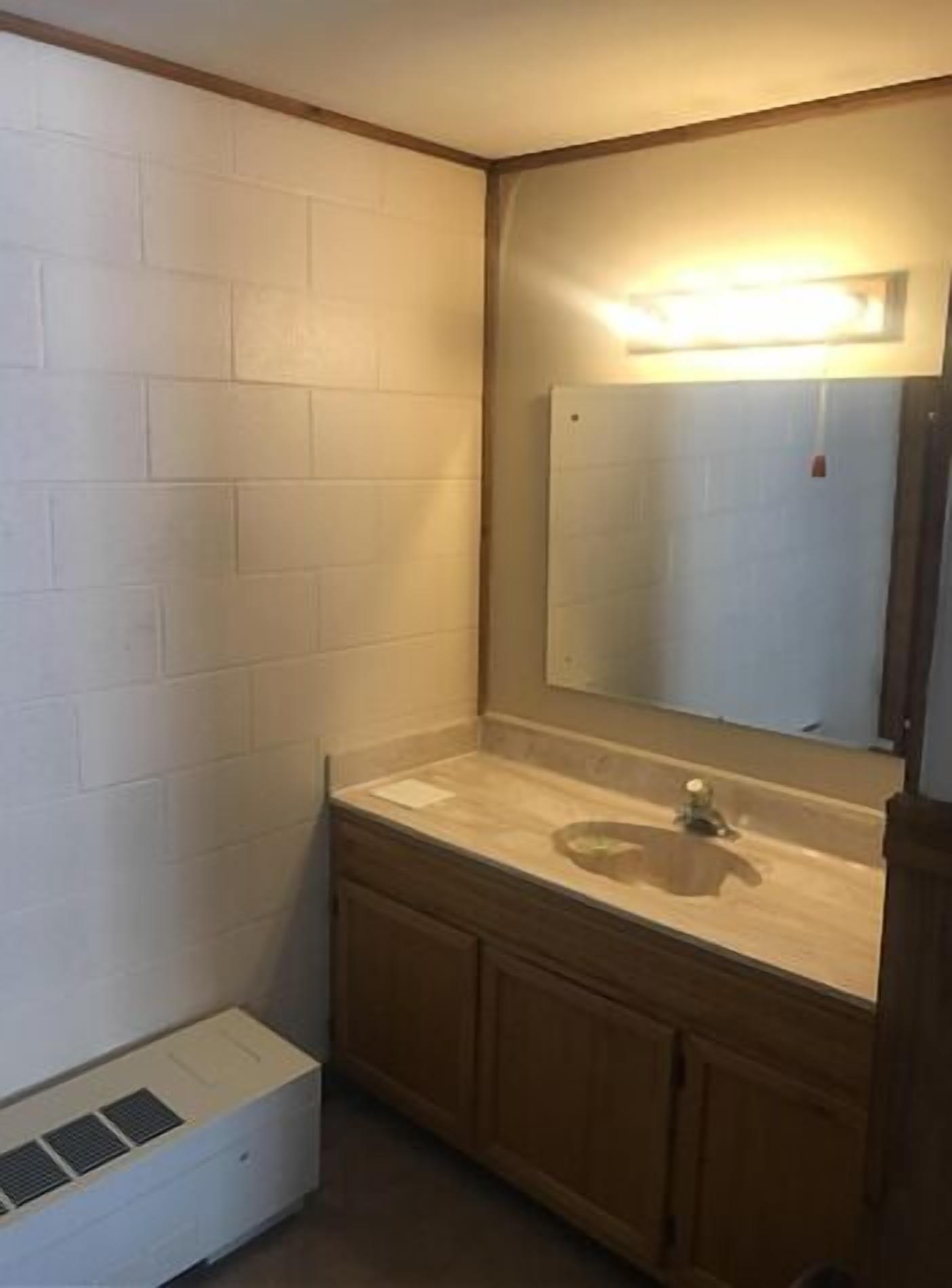 Un baño pequeño con pared de azulejos blancos, tocador de madera, espejo y rejilla de ventilación empotrada.
