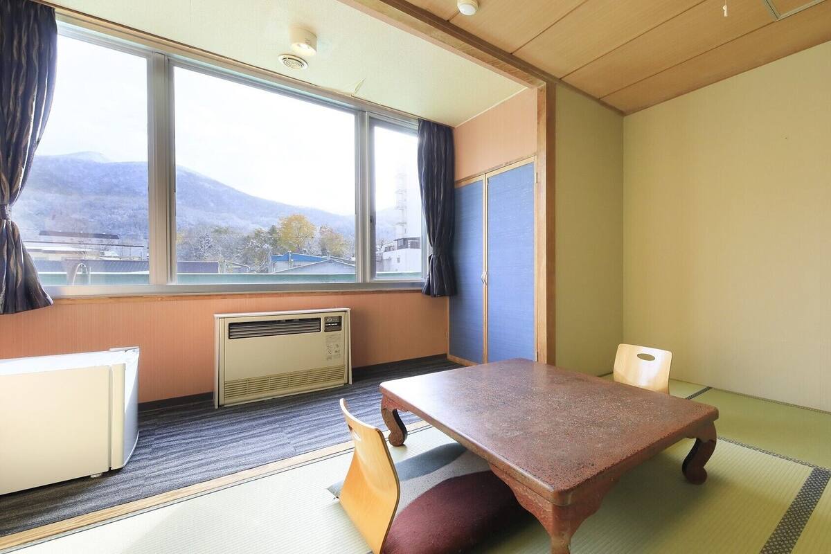 Une pièce avec un sol recouvert de tatamis, une table en bois, un radiateur et une fenêtre donnant sur les montagnes.