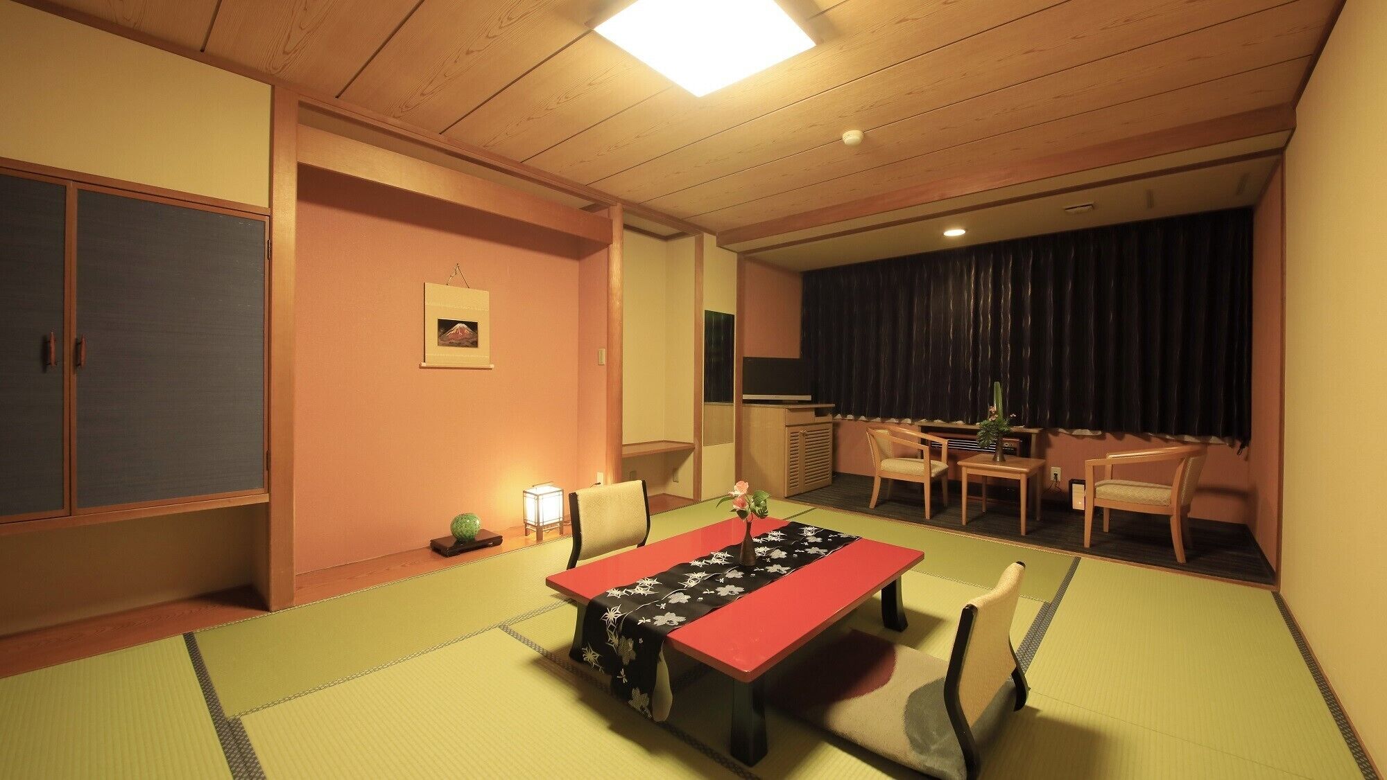 Une pièce de style japonais traditionnel, avec un sol recouvert de tatamis, une table basse, des chaises, un téléviseur et un rideau foncé.